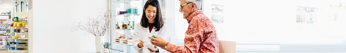 An older gray-haired white man with moustache and glasses being given a prescription by a young female pharmacist